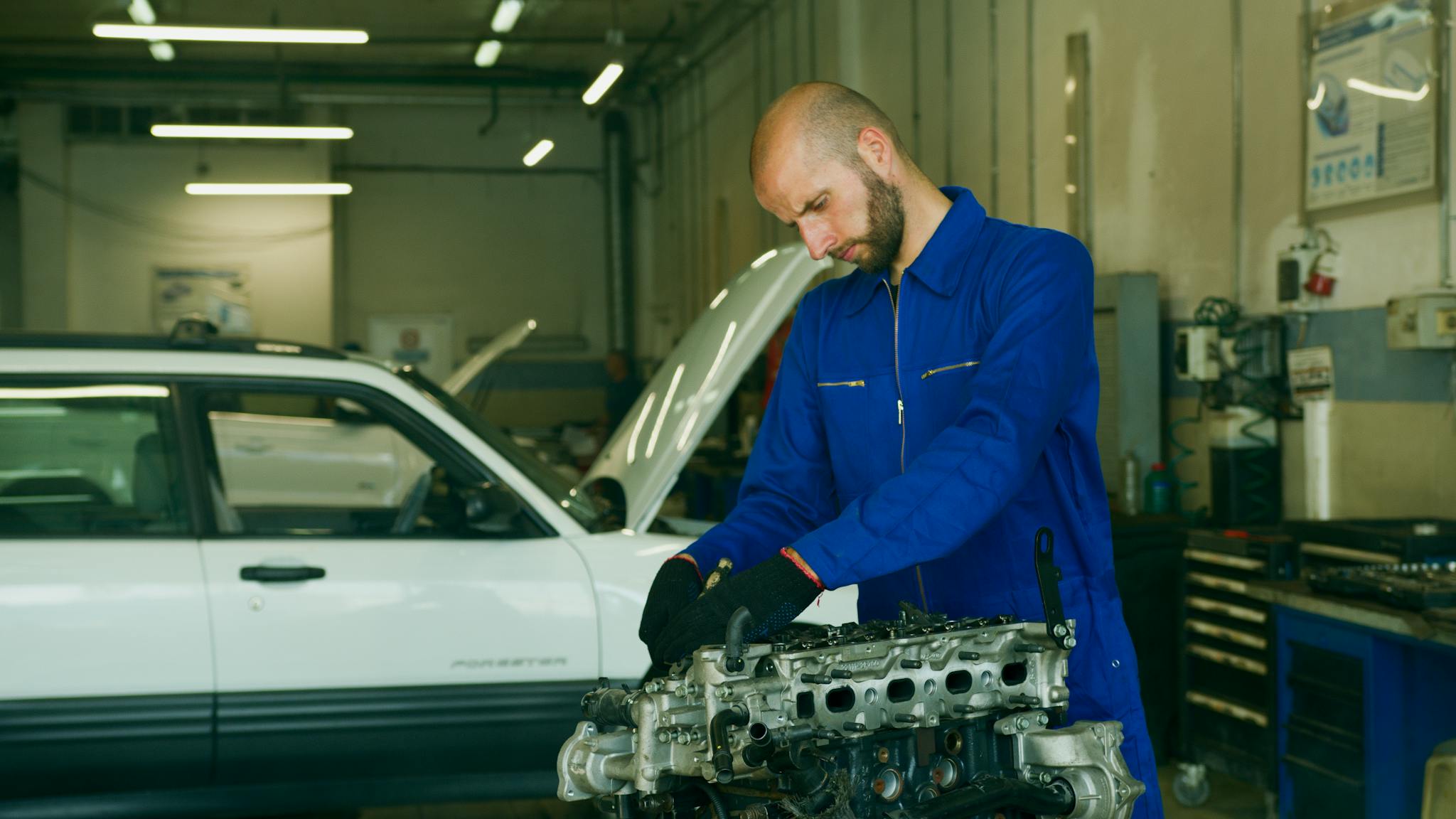 Mechanic in a blue coverall inspecting car engine in a garage workshop.